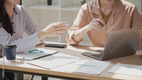 Two young Asian business professionals are sharing a laptop while sitting in a meeting, analyzing financial reports, using a calculator, and discussing data on charts.