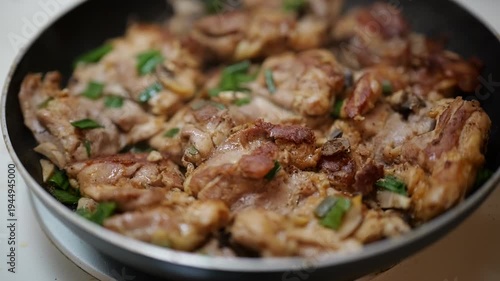 Close-up of seasoned chicken pieces sizzling in a frying pan with garlic and green onions on a stovetop.