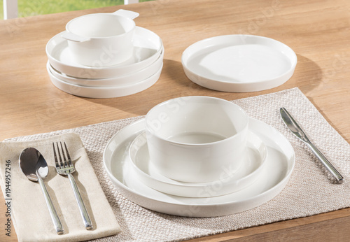 Modern minimalist morning empty dining setup with a pristine white ceramic tableware collection on a wooden table, silver cutlery on a natural fiber placemat, epitomizing a serene breakfast scene.