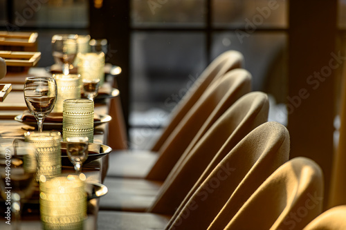 Row of chairs and set table in upscale restaurant. Perspective view of beige chairs and a dinner table with glasses in a warm, lit restaurant hall..