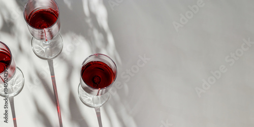 Top view of glasses filled with red wine on white table. Natural sunlight creates long graphic shadows and soft plant leaves silhouettes. Modern minimal summer mood banner, winery aesthetic, copyspace