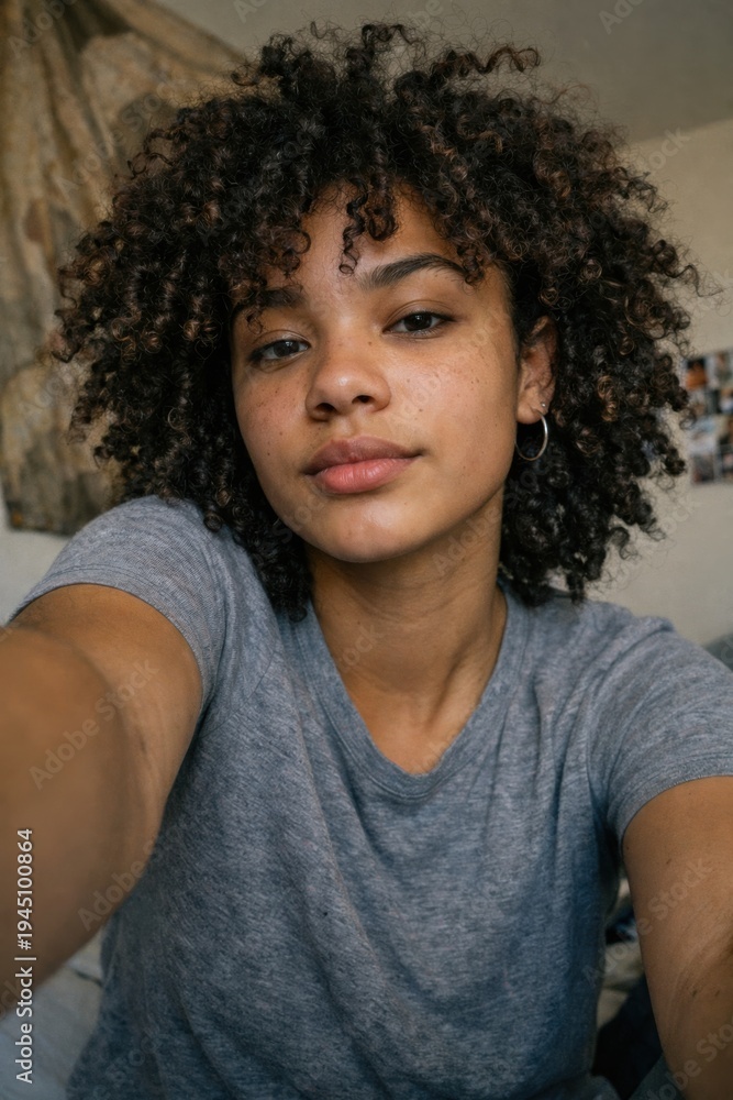 Fototapeta premium Young African American woman with natural curly hair looking thoughtful indoors in casual gray shirt close-up portrait