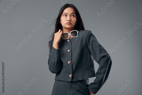 Φωτογραφία Young Asian woman posing in stylish dark gray outfit holding white sunglasses looking away in studio with plain gray background fashion model with long hair and serious expression