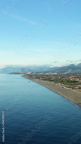 Wide aerial shot of the Fiumicello coastline with calm blue sea, sandy beach, and distant mountains under a clear sky in Italy.
