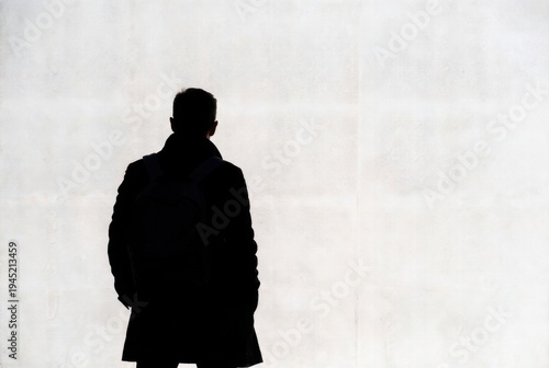 Dark silhouette of a man in a heavy winter coat standing with back to camera against pale wall.