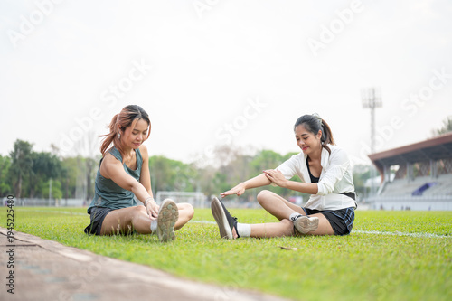 Asian woman and friend stretching arm and leg before exercise on green grass field in sports stadium