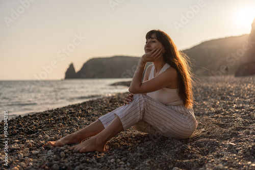 Woman, beach, sunset. Pensive woman sitting on pebble beach, observing the ocean during a peaceful golden hour