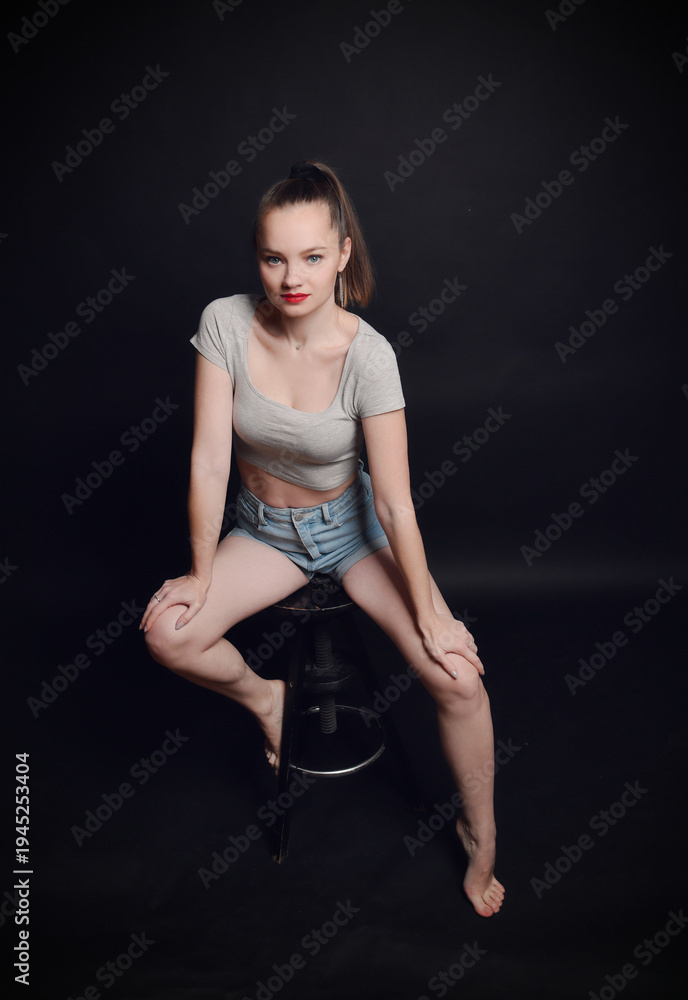Obraz premium Selbstbewusstes Model sitzt auf einem Hocker im Studio und blickt direkt in die Kamera. Modernes Fashion-Porträt mit natürlichem Stil, Konzept für Mode, Lifestyle und Persönlichkeit.