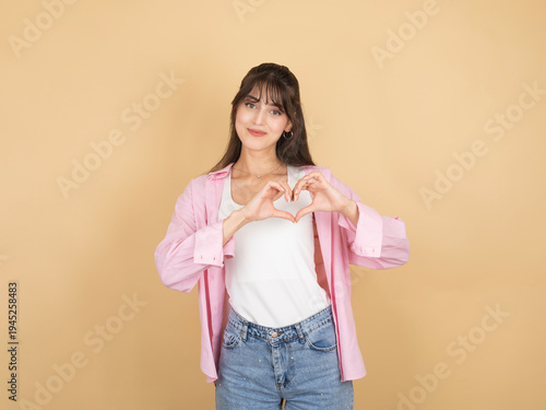 A charming young woman making a heart shape with her hands over her chest while smiling. She is dressed in a pink open shirt and denim jeans against a neutral beige studio background.