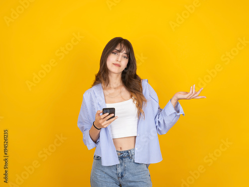 A confused young woman holding a smartphone and gesturing with an open hand against a vibrant yellow background. She wears a striped blue shirt over a white crop top and denim jeans.