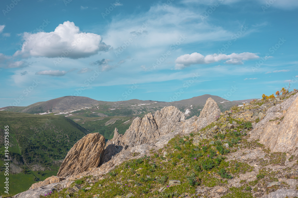 Fototapeta premium Green grasses and rock outcrops on cliff above alpine valley against hills and mountains far away under clouds in blue sky. Sharp rocky outliers on precipice edge in bright sun in cloudy sunny day.