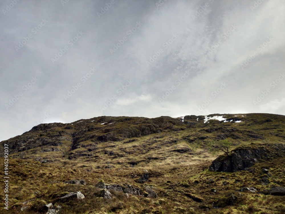 Fototapeta premium Rocky hillside under cloudy sky in Scotland with rugged terrain and sparse vegetation