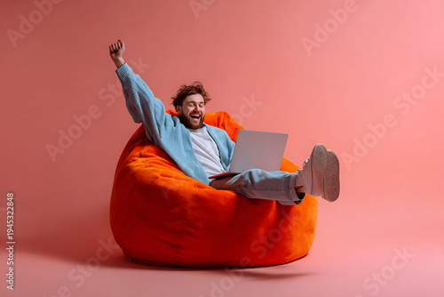 A young man sitting comfortably in an orange bean bag chair with his laptop, expressing excitement and joy.