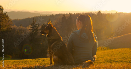 LENS FLARE, CLOSE UP: Woman sits on grassy slope and with her shepherd dog and they both gaze at misty forested hills in golden light. Peaceful moment between owner and pet during an early spring walk