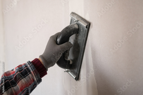 Close-up of worker hand in orange glove sanding plaster on drywall using a plaster hand tool for smooth finish.