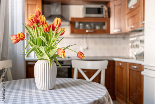 Fresh orange tulips bouquet in white vase on kitchen table with cozy home interior in background. Spring flowers in natural morning light creating warm domestic atmosphere.