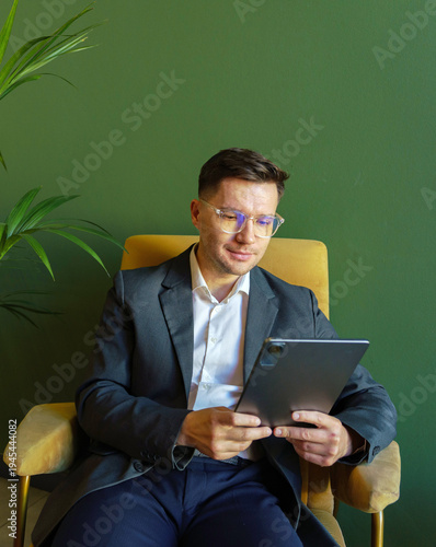 Professional man in eyeglasses sits on a comfortable armchair using a digital tablet against a green wall