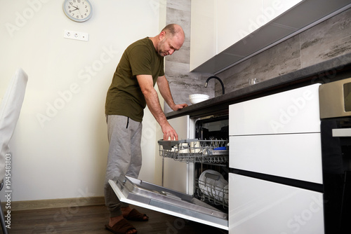 Tableau sur toile A man unloads clean white dishes from the dishwasher in a kitchen with light-colored cabinets and a wood floor