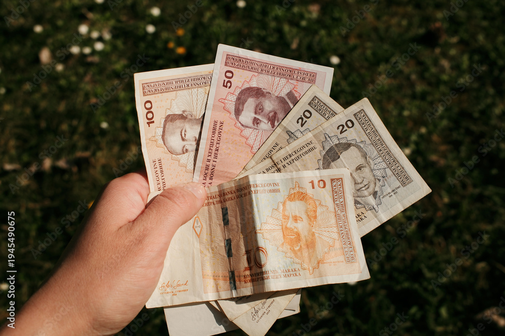 Naklejka premium Hand holding Bosnia and Herzegovina convertible mark banknotes with grass background. Cash currency used for payments and financial transactions in Bosnia and Herzegovina.