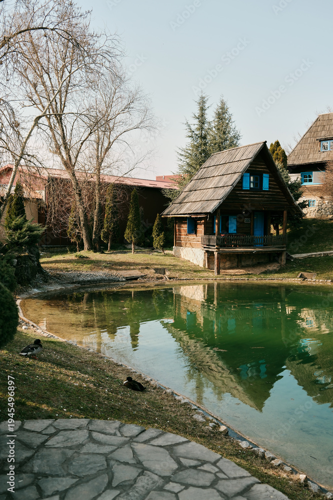 Obraz premium Small wooden rural house with ducks near pond reflection in Stanisici Ethno Village Bijeljina Bosnia and Herzegovina. Concept of countryside architecture cultural tourism and travel destination.