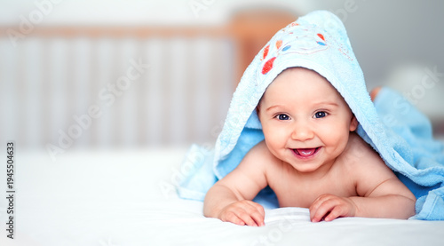 Happy smiling baby boy lying on bed covered with a blue hooded towel. Adorable infant after bath time in a bright nursery setting with copy space for parenting and childcare concepts
