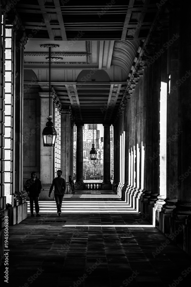 Obraz premium Two people walk through a corridor at Greenwich University. The sunlight creates shadows on the ground and walls. The architecture shows the design of the space.