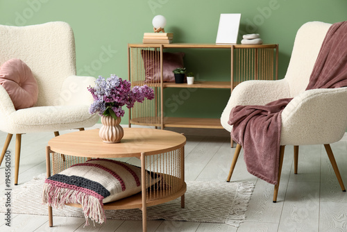Interior of living room with bouquet of lilacs branches on coffee table near white armchairs