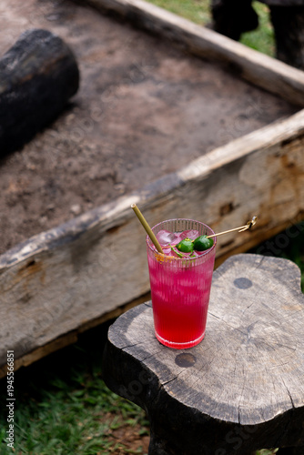 A vibrant pink drink with lime and straws sits on a rustic wooden stump outdoors.