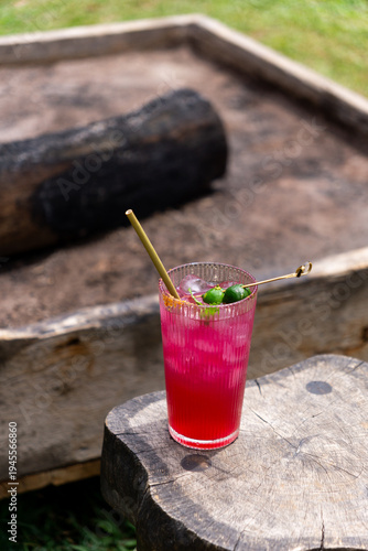 A vibrant pink iced drink with lime and a straw sits on a wooden stool outdoors near a fire pit.