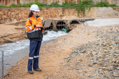 Female environmental scientist in safety gear standing and using laptop and radio for monitoring ecological impact inspection near industrial gas storage tank at fuel terminal site.