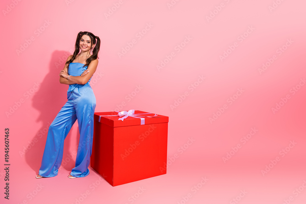 Obraz premium Young chic woman in blue jumpsuit standing beside large red gift box on pink background