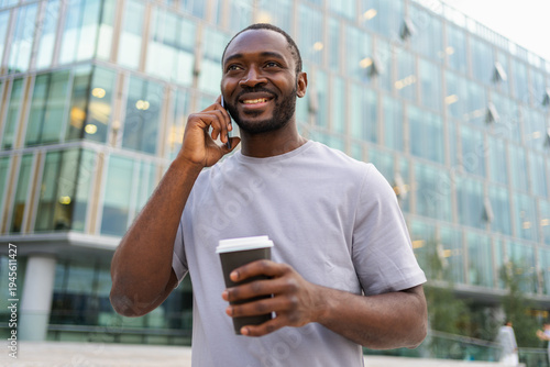 Good morning. African man drinking coffee to go talking on smartphone outdoor. Man with cell phone paper cup walking on street. Man making answering call by cellphone having conversation by mobile