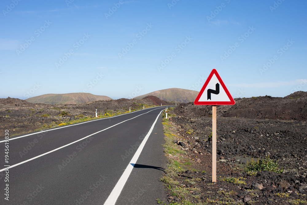 Fototapeta premium A straight, black, endless road with hills and mountains in the background. Spain, Lanzarote. A sign on the side of the road