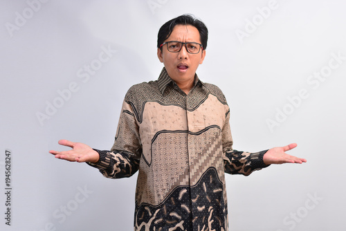 Confused or surprised Indonesian man in batik shirt shrugging his shoulders with open palms