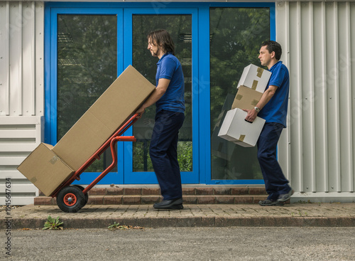 Fotografie Two men delivering large packages on cart in front of warehouse office