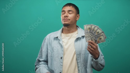 Man holding fanned us dollar bills by his face in studio, gesturing and closing eyes in boastful display; confidence.