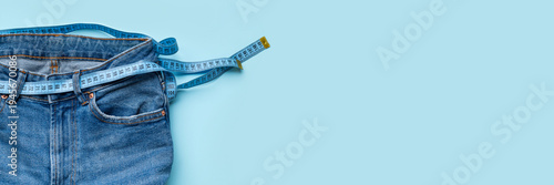 Blue jeans with measuring tape instead of belt on light blue background, flat lay top view. Concepts for weight loss, healthy lifestyle, fitness, excess weight and waist measurement. Long banner