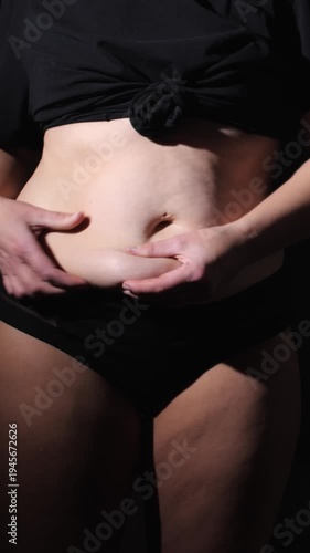Vertical close up of a woman grabbing her belly fat. Concept of body positivity, weight loss, female health and skin texture in dark studio lighting