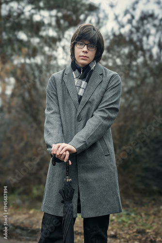 A stylish young man in a gray wool coat and scarf stands outdoors in autumn, holding a closed umbrella