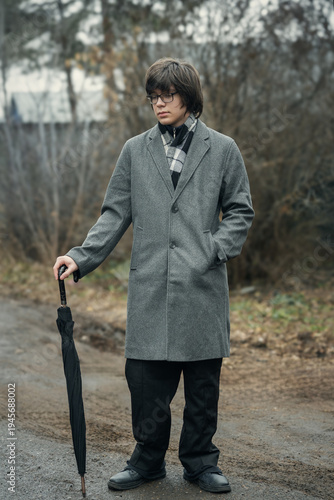 A stylish young man in a gray wool coat and scarf stands outdoors in autumn, holding a closed umbrella