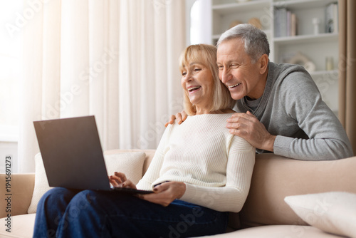 Senior couple sitting together on sofa using laptop and smiling warmly at home. Mature man and woman enjoying online activity representing family lifestyle, technology use and retirement comfort