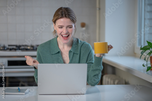 Fotografie Excited young woman sitting at kitchen table with laptop, celebrating good news,