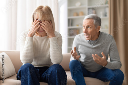Senior woman sitting on sofa covering face with hands while her husband speaks emotionally beside her. Mature couple experiencing relationship stress and difficult conversation at home.