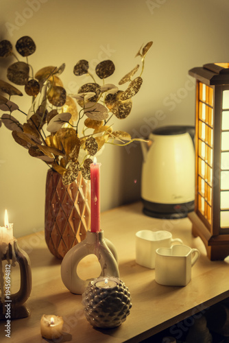 A cozy evening scene featuring a wooden table with glowing candles, a decorative vase with gold foliage, heart-shaped mugs, and an elegant Japanese-style wooden lamp, creating a warm atmosphere.