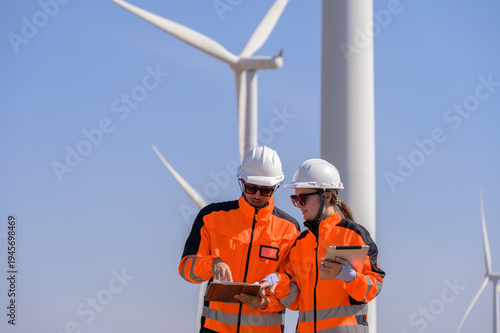 Two wind energy engineers wearing safety helmets and high visibility jackets review technical data at wind farm for inspection and maintenance planning