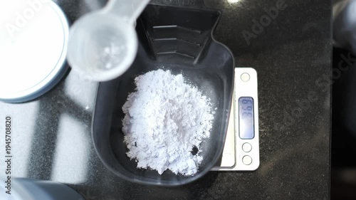 Hairstylist pouring white bleaching powder from a plastic scoop into a black mixing bowl on a digital scale. Top view