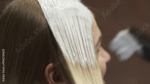 Professional hair stylist applying white bleach or hair dye to a section of blonde hair using a brush in a salon