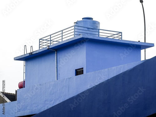 Fotografija A minimalist shot of a bright blue industrial rooftop featuring a large water storage tank, safety railings, and clean geometric lines against a pale sky