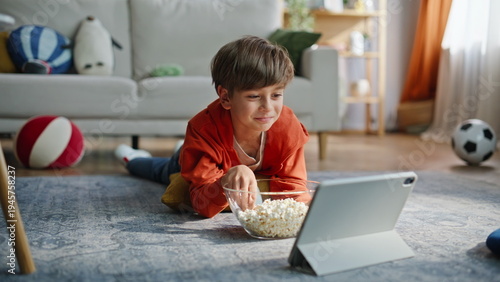 Smiling boy watching tablet cartoon eating popcorn lying apartment floor closeup