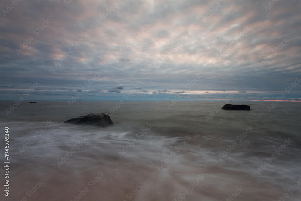 Fototapeta premium A tranquil Baltic Sea showing smooth, long-exposure waves flowing around large coastal rocks on a sandy beach beneath a pastel cloudy sunset, conveying calm, solitude and natural coastal beauty. 